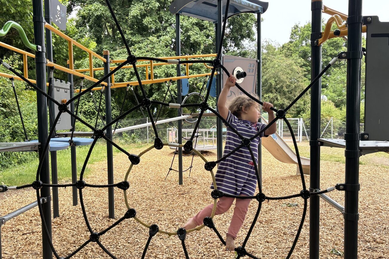 Ein Kinderspielplatz mit Klettergerüst, ein Mädchen in lila-weiß gestreiftem Kleid und rosafarbener Hose klettert.