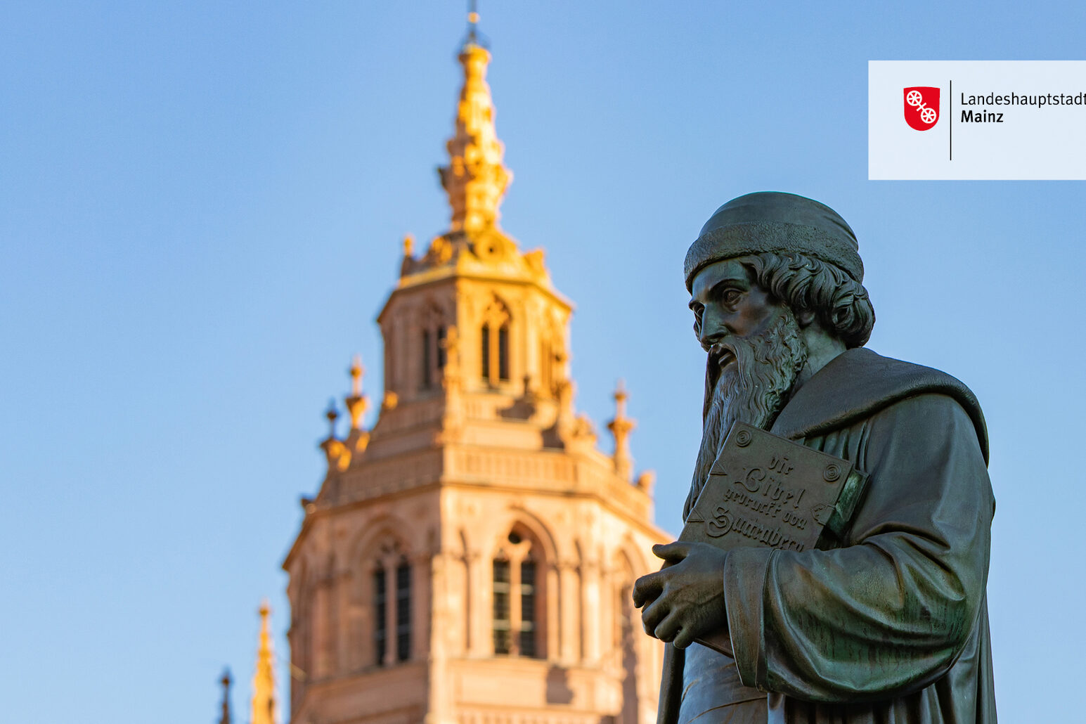 Spitze des Westturms des Mainzer Doms im Sonnenschein, daneben die Statue von Johannes Gutenberg mit Bibel unterm Arm, im Hintergrund blauer Himmel.