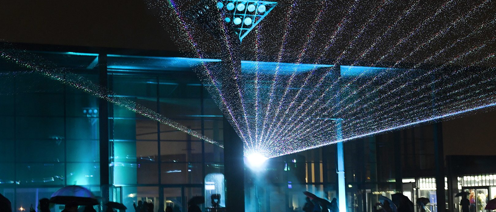 Illumination eines Mainzer Gebäudes, bei Dunkelheit, vor dem Menschen mit Regenschirmen stehen.