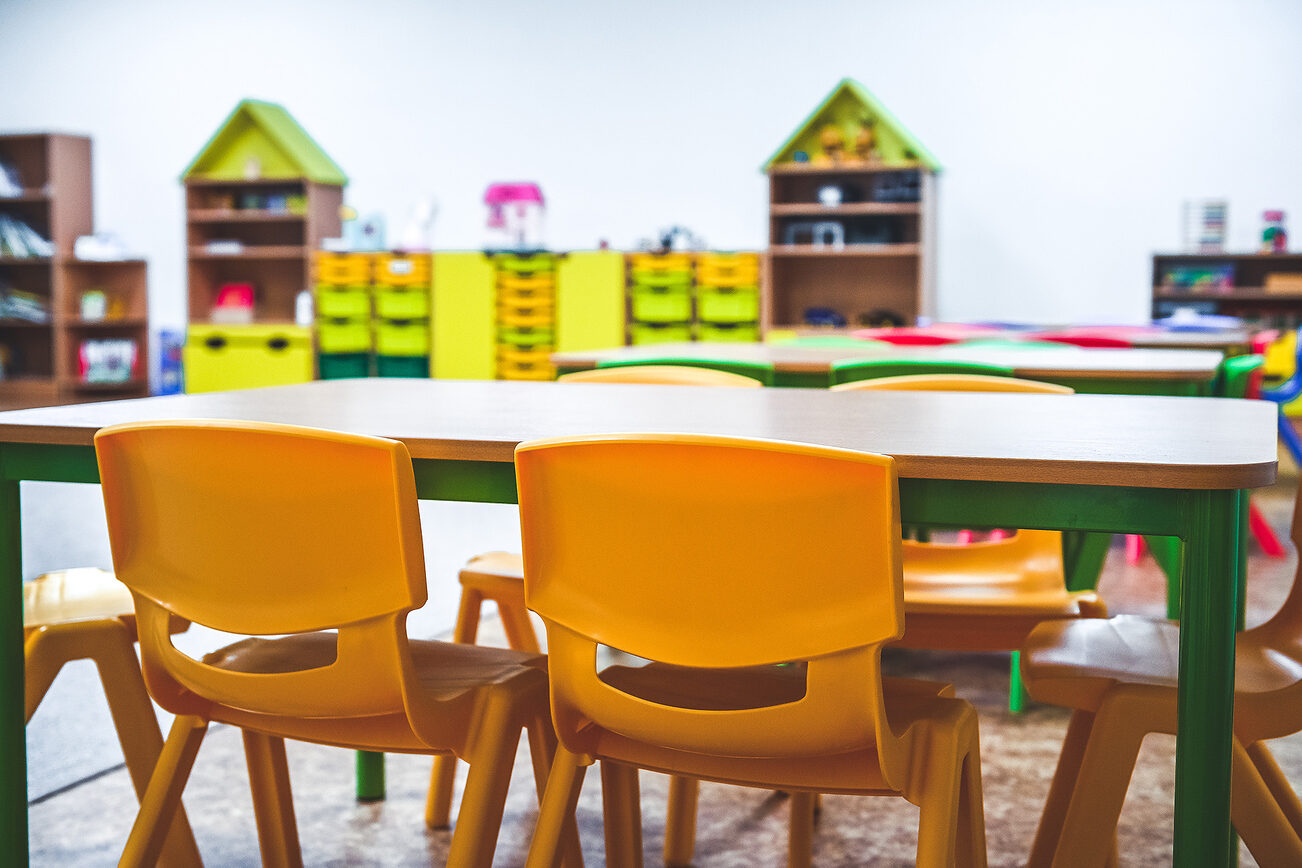 Gelbe Plastikstühle an einem Tisch im Vordergrund in einer Kindertagesstätte, im Hintergrund Regale mit glben und grünen Fächern.
