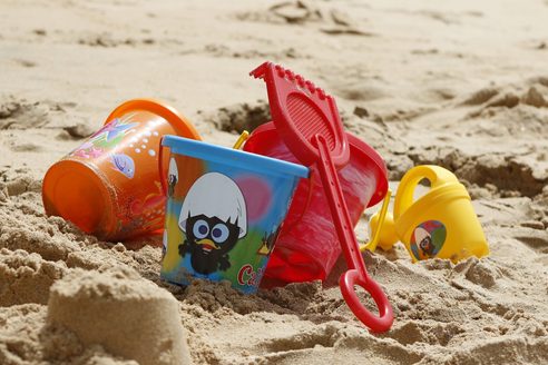 3 bunte Spielzeug-Eimer, eine rote Kinderschaufel und eine Kindergießkanne liegen im weichen Sand.